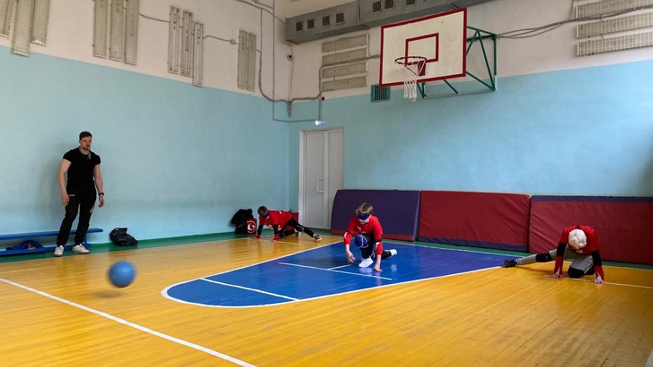 Три школьника в спортивной форме с повязками на глазах лёжа на полу спортзала играют в голбол, у стены зала стоит тренер-судья