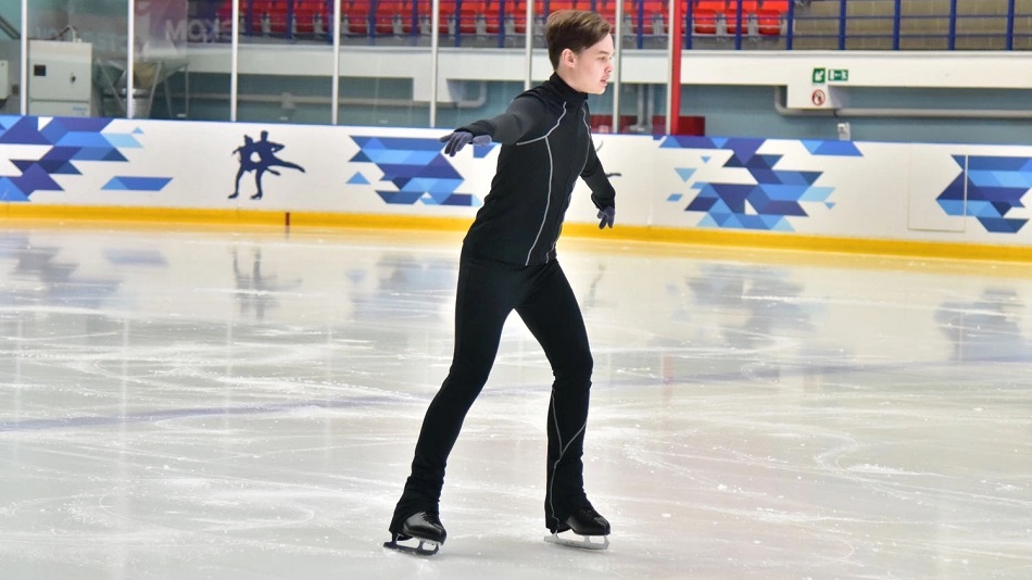 Юноша с аутизмом на льду выступает в одиночном фигурном катании (в полный рост, проезд слева направо мимо камеры)