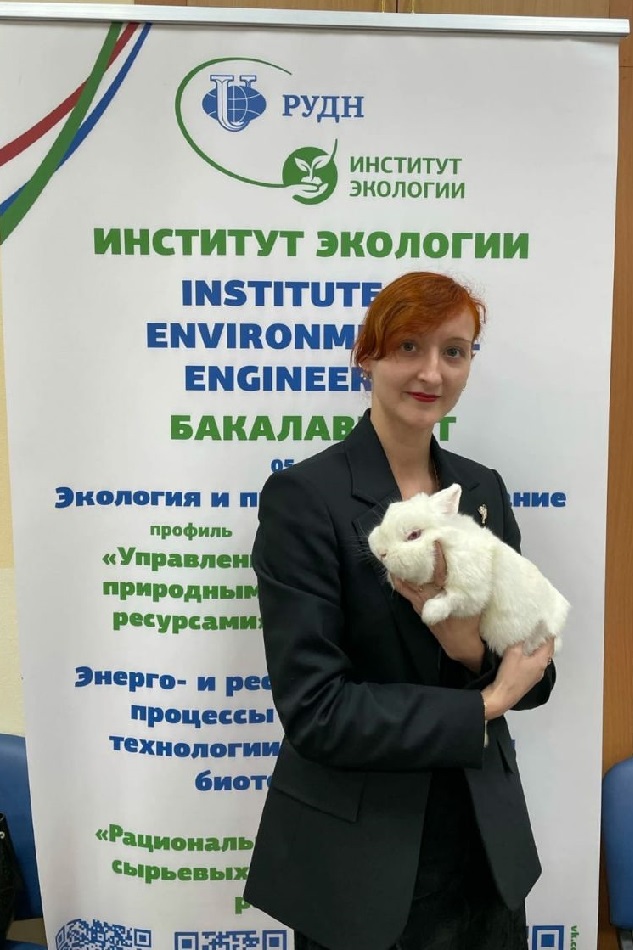 Ирина Головачёва с маленькой белой собачкой на руках стоит перед стендом Инстита экологии Университета дружбы народов