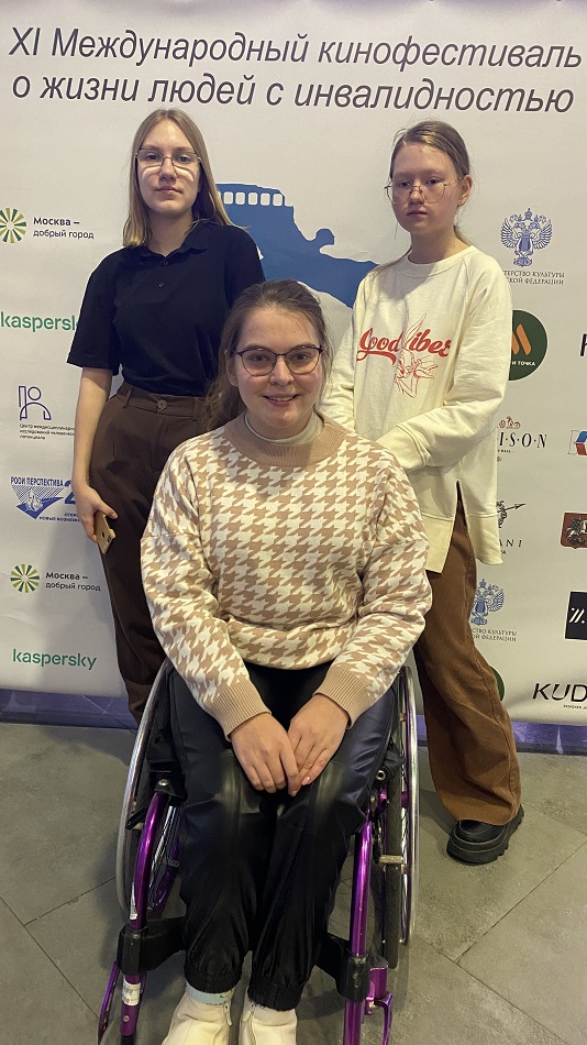 Ксения Каминская в инвалидной коляске с двумя друзьями на фоне стенда 11 кинофестиваля "Кино без барьеров"